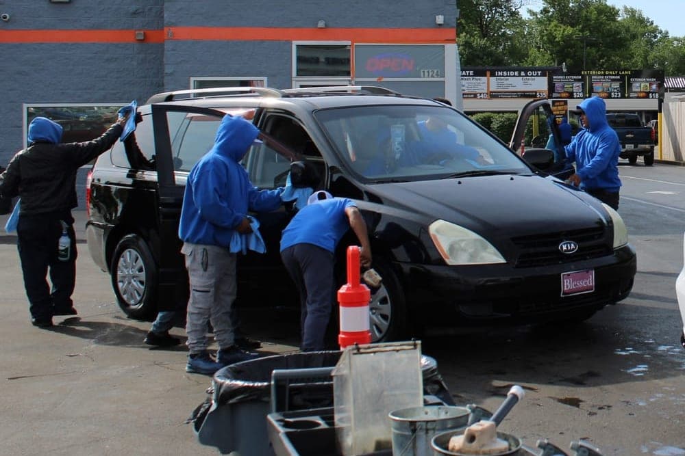 The Lexington Auto Spa crew cleaning the interior of a black Kia minivan with its doors open, one member reaching inside while another wipes down the glass